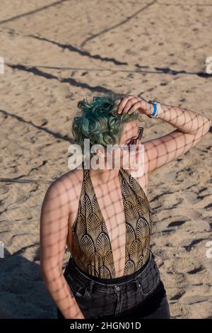 Jeune femme avec des cheveux bleus verts sur le sable de la plage à l'ombre de la grille. Banque D'Images