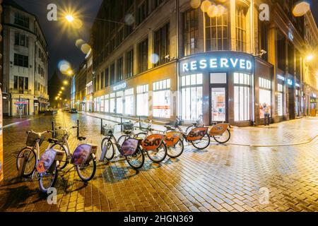 Riga, Lettonie.Rangée de bicyclettes à louer au parking à vélos près de la boutique de vêtements le réservé sur la rue Valnu dans l'éclairage de nuit.Réservé est Banque D'Images