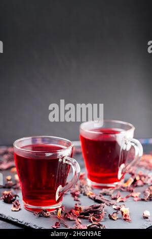 Thé à l'hibiscus rouge dans une tasse en verre sur fond sombre.Tasse de thé hibiscus et pétales d'hibiscus secs sur un plateau en ardoise.Copier l'espace Banque D'Images