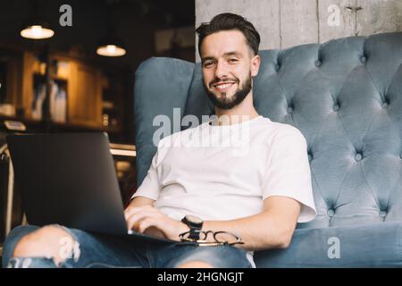 Portrait d'un homme hipster qualifié souriant à l'appareil photo tout en faisant un travail à distance d'ordinateur portable dans l'espace de travail de collègue Banque D'Images