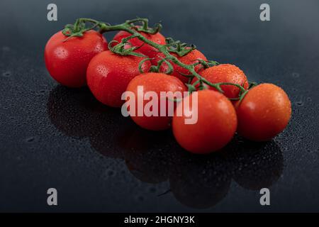 Belles tomates de vigne fraîches (cerise) avec des gouttes d'eau, brutes, entières et coupées en deux. Sur fond d'ardoise texturé noir. Ingrédients de la Méditerranée Banque D'Images