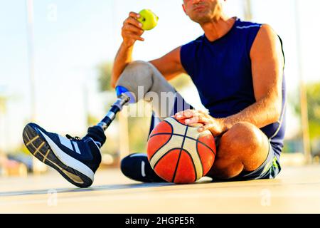 homme sportif handicapé dans les vêtements de sport et avec joueur de basket-ball de jambe artificielle au coucher du soleil manger une pomme Banque D'Images