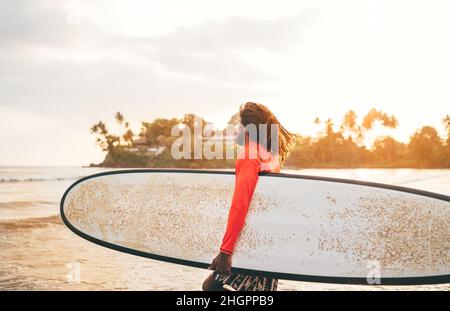 Adolescent noir à poils longs avec planche de surf cirée prête pour le surf avec rétroéclairage au coucher du soleil.Il marche dans les vagues de l'océan Indien.Sports nautiques extrêmes an Banque D'Images