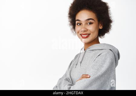 Portrait d'une femme noire souriante avec cheveux afro, bras croisés sur la poitrine, port de sweat à capuche gris sportif, air heureux et confiant, fond blanc Banque D'Images