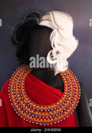 Un mannequin en plastique avec une perruque noire et blanche, une robe d'emballage rouge et un collier à perles Maasai aux couleurs vives. Banque D'Images