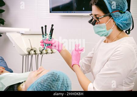 Vue latérale de la femme dentiste dans les jumelles en loupe dentaire, les gants médicaux et les outils de maintien de masque pour guérir les dents du patient dans la clinique dentaire. Banque D'Images