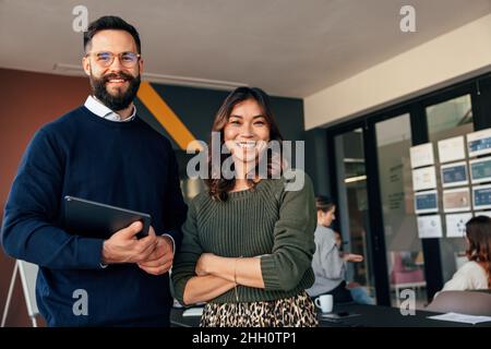 Deux entrepreneurs heureux souriant à la caméra joyeusement.Jeunes hommes d'affaires debout dans une salle de réunion avec leurs collègues en arrière-plan.Plongeurs Banque D'Images