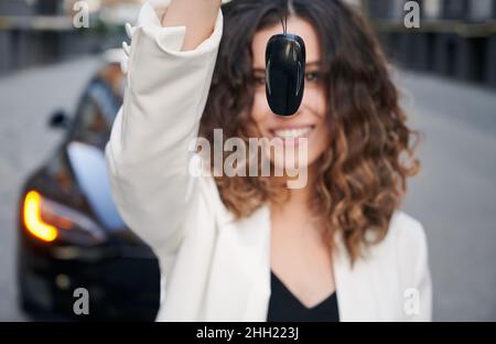 Portrait d'une femme de vente floue tenant la clé électronique de voiture comme publicité d'entreprise, courtier caucasien réussi montrant la télécommande pour le véhicule noir de compagnie appréciant le mode de vie d'affaires Banque D'Images