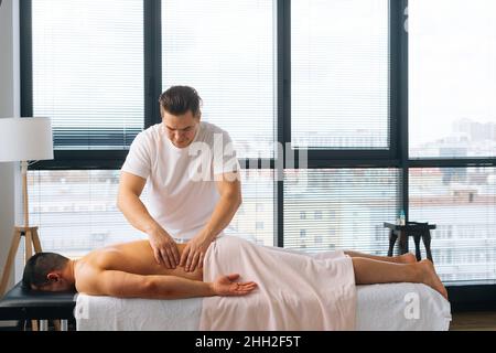 Prise de vue large d'un masseur masculin professionnel avec des mains fortes, massant le côté du dos à un sportif musclé allongé sur l'estomac, Banque D'Images