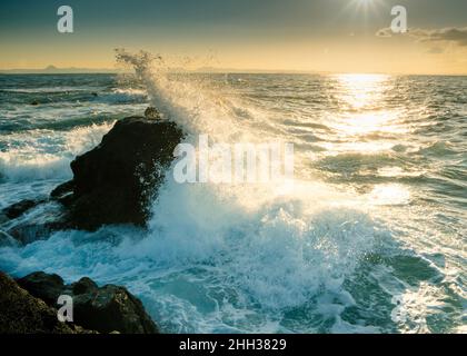 Tandis que les vagues froides, sombres et bleues d'hiver s'écrasont contre les rochers, le soleil levant baigne la froideur avec un soleil doux, chaud et doré. Banque D'Images
