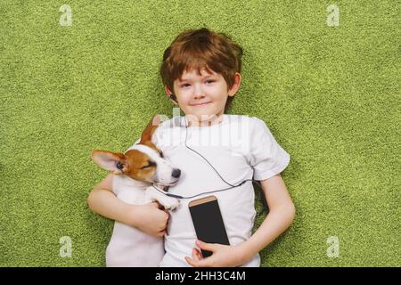 Un petit garçon avec un chien écoute de la musique au téléphone tout en étant allongé sur la moquette.Amitié, concept de séjour à la maison. Banque D'Images