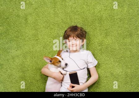 Un petit garçon avec un chien écoute de la musique au téléphone tout en étant allongé sur la moquette.Amitié, concept de séjour à la maison. Banque D'Images