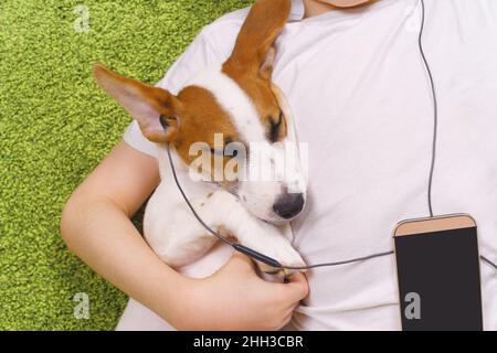 Un petit garçon avec un chien écoute de la musique au téléphone tout en étant allongé sur la moquette.Amitié, concept de séjour à la maison. Banque D'Images