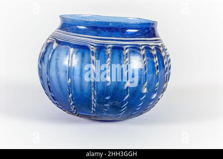 Bol côtelé en verre mi-1st Century A.D. Roman bleu cobalt translucide ; traînée en blanc opaque. Rebord rodé et inégal. Col concave court. Corps globulaire profond courbé à fond plat. Plaie striée spiralée autour du corps, avec grande surface circulaire sur le fond.Côtés en vingt-trois nervures verticales irrégulières. Brisés avec de grandes fissures autour du corps et une petite puce dans le rebord; quelques bulles; morcellement, piqûres, faible iridesence,et l'altération de la traînée autour du bas du corps et entre les nervures.Bol en verre côtelé.Romain. Mi-1st siècle A.D..Verre ; soufflé, traîné et outillaté.Début de l'Impériale.Verre Banque D'Images