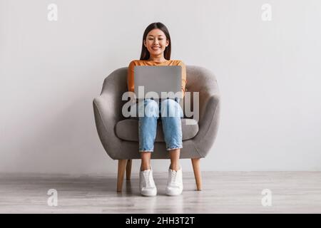 Jolie jeune femme asiatique assise dans un fauteuil, travaillant sur un ordinateur portable contre un mur blanc de studio, pleine longueur Banque D'Images