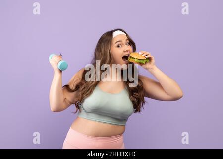 Gai drôle faim jolie millénaire plus grande taille jeune femme en sport uniforme manger le hamburger et de levage haltères Banque D'Images