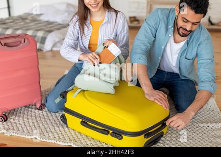 Les jeunes conjoints emballant les vêtements dans une valise et la femme mettant le passeport avec les billets dans le sac, se préparant pour les vacances Banque D'Images