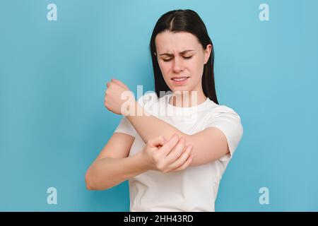 Malheureux triste jeune femme caucasienne 20s ans ayant des douleurs dans son coude, se réchauffant la main blessée après un traumatisme, posant isolé sur le dos bleu de couleur Banque D'Images