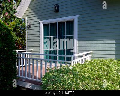 Fenêtre blanche de maison avec verre, rideau vert sur la terrasse vide le petit bois vert clair résident sur le jardin vert, vue de l'extérieur. Banque D'Images