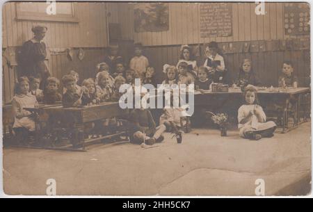 Les enfants assis à leur bureau dans une salle de classe, certains ont des jouets et des fleurs devant eux, d'autres font de la broderie, un enseignant est debout d'un côté.La comptine Jack et Jill est écrite sur le mur.Photographie prise au début du 20th siècle Banque D'Images