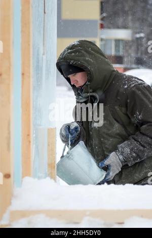 Un constructeur de ville de glace dans une veste à capuchon verte verse l'eau d'un seau au-dessus d'un bloc de glace Banque D'Images