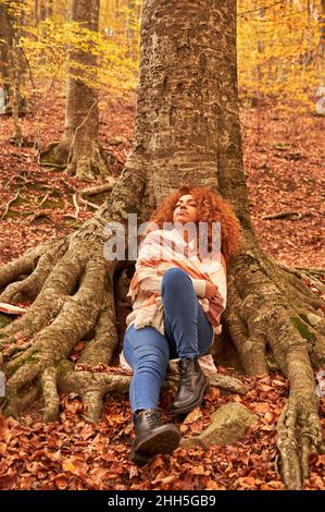 Jeune femme se détendant par arbre dans la forêt d'automne Banque D'Images