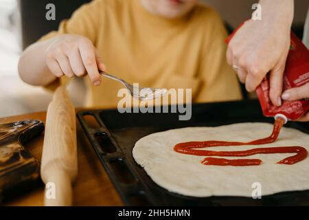 Mère et fils étalant la sauce tomate sur la pâte dans le plateau à la maison Banque D'Images