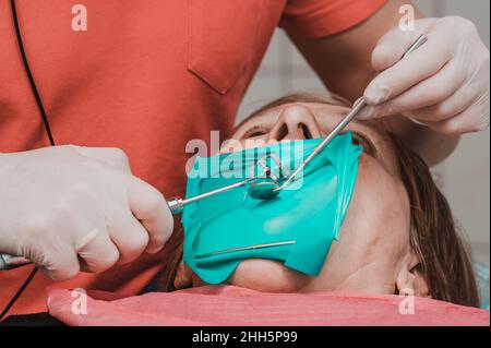 Une patiente avec un écarteur de joue avec un cercueil vert dans une clinique dentaire, un dentiste en gants de latex examine ses dents avec un bore et un miroir, a Banque D'Images