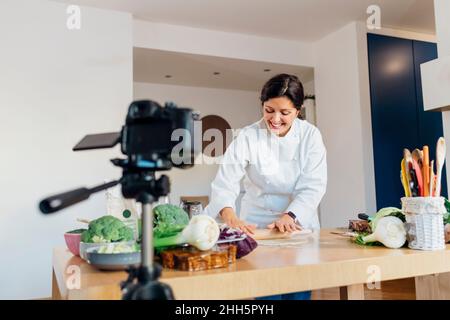 Un chef souriant fait des conseils sur la pâte à rouler à l'aide d'un appareil photo dans la cuisine à la maison Banque D'Images