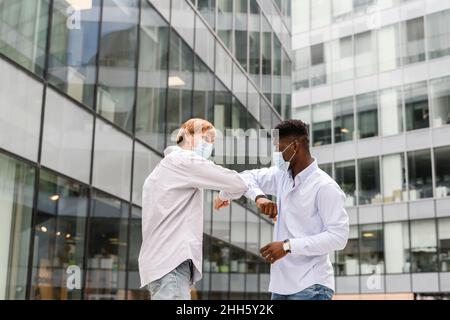 Amis dans les masques chirurgicaux faisant le coude de choc par le bâtiment moderne Banque D'Images