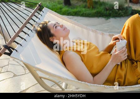 Femme contemplative avec livre et téléphone mobile couché dans hamac Banque D'Images
