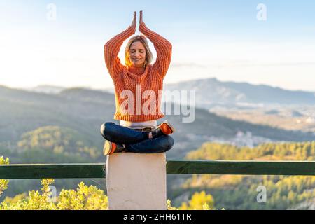 Femme souriante méditant avec les yeux fermés et les mains soulevées sur la main courante au parc Banque D'Images
