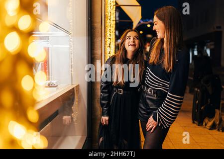 De jeunes amis souriants se tenant près de la fenêtre illuminée du magasin la nuit Banque D'Images