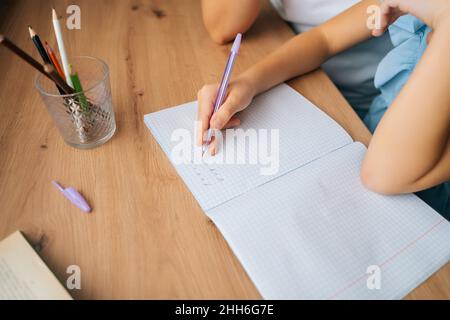 Gros plan rogné de fille primaire faisant des devoirs écrivant dans un livre avec un stylo avec une jeune mère assis à la table de la maison. Banque D'Images