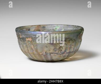 Bol en verre nervuré fin du 1st siècle C.-B.–début du 1st siècle A.D. Roman bleu pâle translucide vert.rebord vertical arrondi; côtés profonds avec bande lisse autour du haut, courbé vers le bas légèrement concave.à l'intérieur, une large rainure horizontale sous le rebord et deux étroites rainures étroites découpées dans une bande autour du bas du corps;Sur l'extérieur, trente-huit nervures peu profondes et légèrement obliques, s'inclinant vers le bas et s'étendant jusqu'au bord du fond. Intactes, mais certaines fissures internes; bulles de piqûre; morcellement, piqûres profondes et iridescences,Avec des plaques d'altération crémeuse épaisse. Marques de meulage rotatives à l'intérieur et sur Banque D'Images