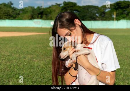 Jeune femme aux cheveux bruns droits et lâches, tenant un petit chien demi-race avec les deux mains près de sa poitrine, son visage appuyé sur la tête du chien, Banque D'Images