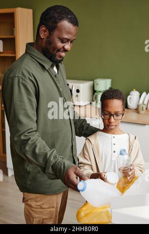 Portrait vertical du père et du fils afro-américains heureux mettant du plastique dans des bacs de recyclage à la maison Banque D'Images