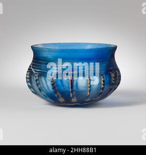 Bol côtelé en verre mi-1st Century A.D. Roman bleu translucide ; traînée en blanc opaque. Rebord extérieur, avec rebord fendu et rodé ; col concave court ; corps globulaire incurvé dans un fond convexe peu profond. Traînée appliquée comme un gros blob et enroulée en spirale sur le fond, puis d'un côté à l'autre du cou.Le côté a été taillé dans dix-sept côtes irrégulières, verticales ou obliques.intactes, mais certaines parties de la traînée ont disparu à travers les intempéries; de nombreuses bulles, certaines grandes; piqûres de bulles de surface, de juxtaposition, d'iridescenence, et des taches de crémeux intempéries, en particulier autour des côtes.bol bleu vif avec décoration de fil blanc, nervure verticale Banque D'Images
