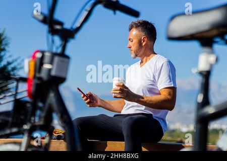 bel homme en t-shirt blanc assis sur le banc à côté du scooter écologique avec belle vue sur les collines et boire un café à emporter Banque D'Images