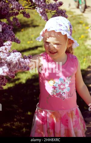 Belle petite fille blonde avec panama sur sa tête jouant dans le jardin de printemps avec le Bush en fleur de lilas pourpre. Banque D'Images