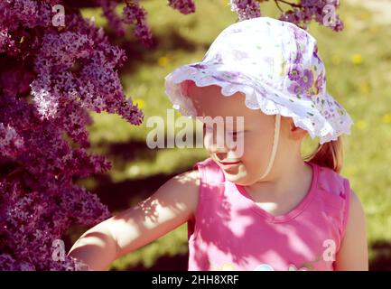 Belle petite fille blonde avec panama sur sa tête jouant dans le jardin de printemps avec le Bush en fleur de lilas pourpre. Banque D'Images