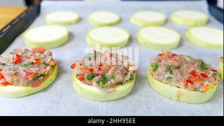 Courgettes farcies de viande hachée et de légumes crus sur une plaque à pâtisserie. Les mains des femmes répartissent la viande hachée sur des cercles de courgettes. Banque D'Images