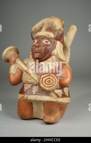 Bouteille à bec en étrier avec Warrior Figure 1st–4th Century Moche ce bateau en céramique en forme de guerrier au genou incliné a été fabriqué par des artisans de la culture Moche de la côte nord du Pérou.La figure porte des régales de combat, y compris un casque conique fixé sous le menton et doté d'un croissant en métal projetant vers le haut, une tunique rouge et blanche, une ceinture, un collier avec de longues perles en forme de liège de champagne, des ornements de poignet et un tissu de longe.Il porte une mace et un petit bouclier avec un dessin de cercles concentriques blancs et rouges.Le pigment plus foncé de son visage indique qu'il porte fac Banque D'Images