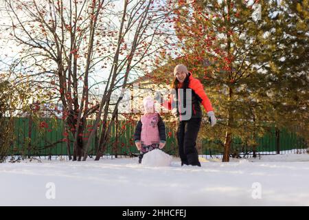 Jolie petite fille et mère roulant de grandes boules de neige pour construire bonhomme de neige souriant sur cour en soirée avec rowan et sapins en arrière-plan Banque D'Images