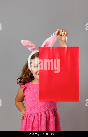 Pâques.Une petite fille gaie dans une robe rose sort de derrière un sac de shopping.Enfant mignon sur fond gris isolé. Banque D'Images