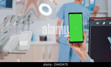 Gros plan du dentiste tenant un smartphone avec écran vert pour le soin de la dentisterie.Spécialiste de la dentisterie regardant la clé chromique et le modèle de maquette isolé sur le téléphone mobile. Banque D'Images