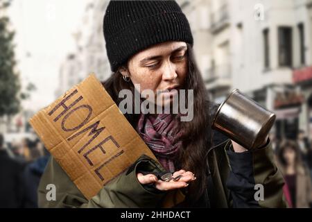 Une femme mendiante tient une pancarte en carton avec l'inscription sans-abri et regarde les pièces d'une tasse de mendicité en acier.Arrière-plan non focalisé avec rue et Banque D'Images