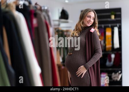 Jeune femme blonde enceinte essayant de porter des vêtements dans un magasin de vêtements Banque D'Images