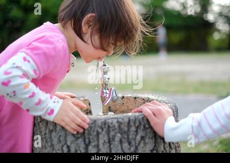 Petite fille aux cheveux bruns courts qui boit de l'eau provenant d'une fontaine dans le parc.Printemps. Banque D'Images