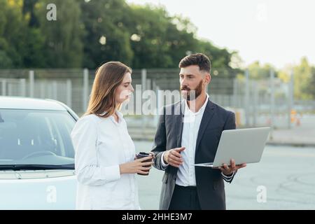 Un vendeur de voitures, recommande une voiture à une femme acheteur, inspecte les voitures dans la salle d'exposition, le vendeur utilise un ordinateur portable pour visualiser les informations techniques sur la voiture Banque D'Images